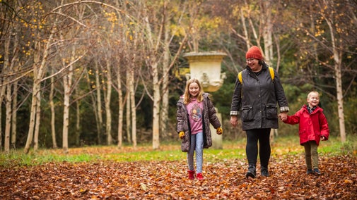 A woman and two children, dressed in warm coats and bobble hats, walking through fallen leaves with autumn trees and a garden statue in the background
